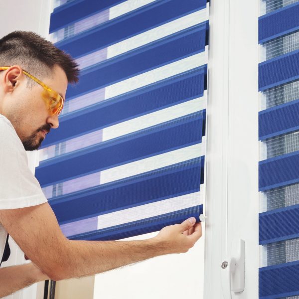 Male builder carpenter pulling down a blue jalousie on a window. Repairing and inspection fabric curtains. Duo system day and night blind. Interior decor element Male builder carpenter pulling down a blue jalousie on a window. Repairing and inspection fabric curtains. Duo system day and night blind. Interior decor element.